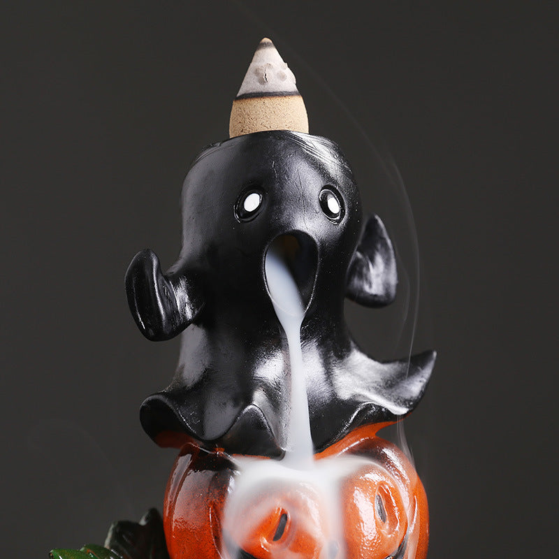 Black ceramic ghost incense burner on top of an orange pumpkin, emitting smoke from its open mouth. A cone of incense is placed on its head.