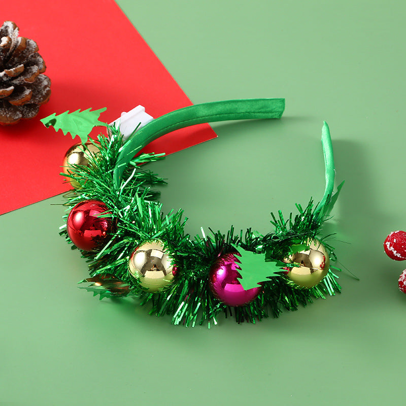 Festive green headband decorated with tinsel, red, gold, and purple ornaments, and green leaves, lying on a green surface next to a red card and pinecone.