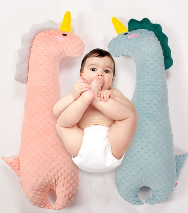 Baby in a diaper lies between two plush dinosaur pillows, one pink and the other blue. The baby is sucking on their toes while looking upwards.