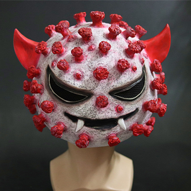 A mask resembling a virus particle with a sinister face, featuring red horns, spikes, and pointed teeth, displayed on a mannequin head against a dark background.