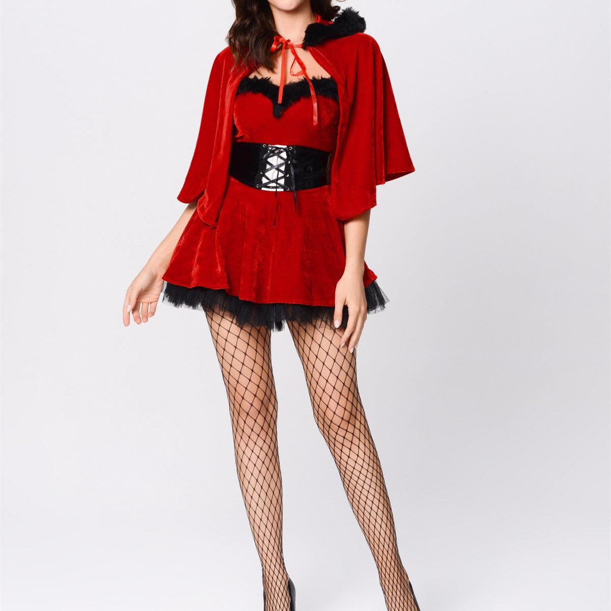 A woman in a red velvet cape and dress with black lace trim and a black corset belt, paired with fishnet stockings and heels, standing against a plain background.
