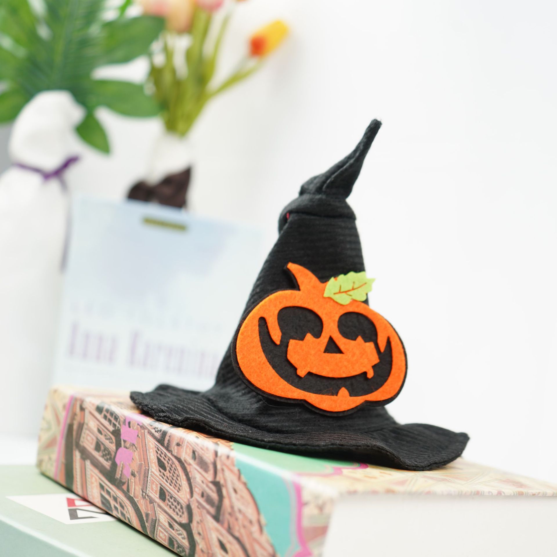 A black witch hat with a smiling orange jack-o'-lantern decoration sits atop two books. Blurred background features flowers and abstract decor.