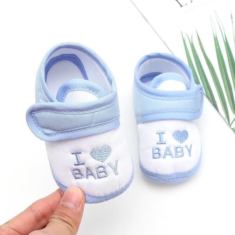 Pale blue and white baby shoes with "I ♥ BABY" embroidered on them. A hand holds one shoe. A leaf and paper are partially visible in the background.