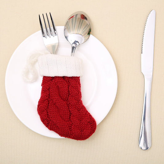 A red knitted Christmas stocking holds a fork and spoon on a white plate. A knife is placed on the right, all set against a beige background.