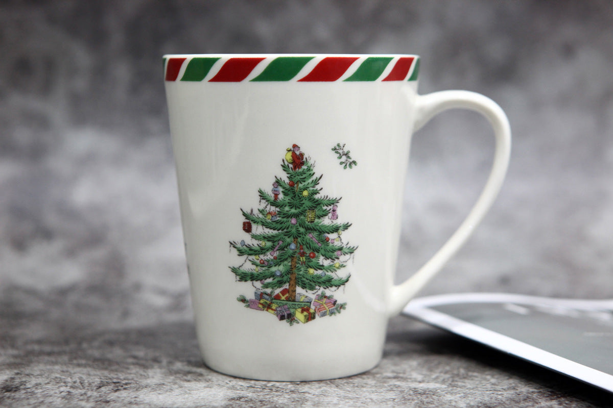 White mug with a festive Christmas tree design, adorned with presents and decorations. Red and green striped rim. Set against a neutral textured background.