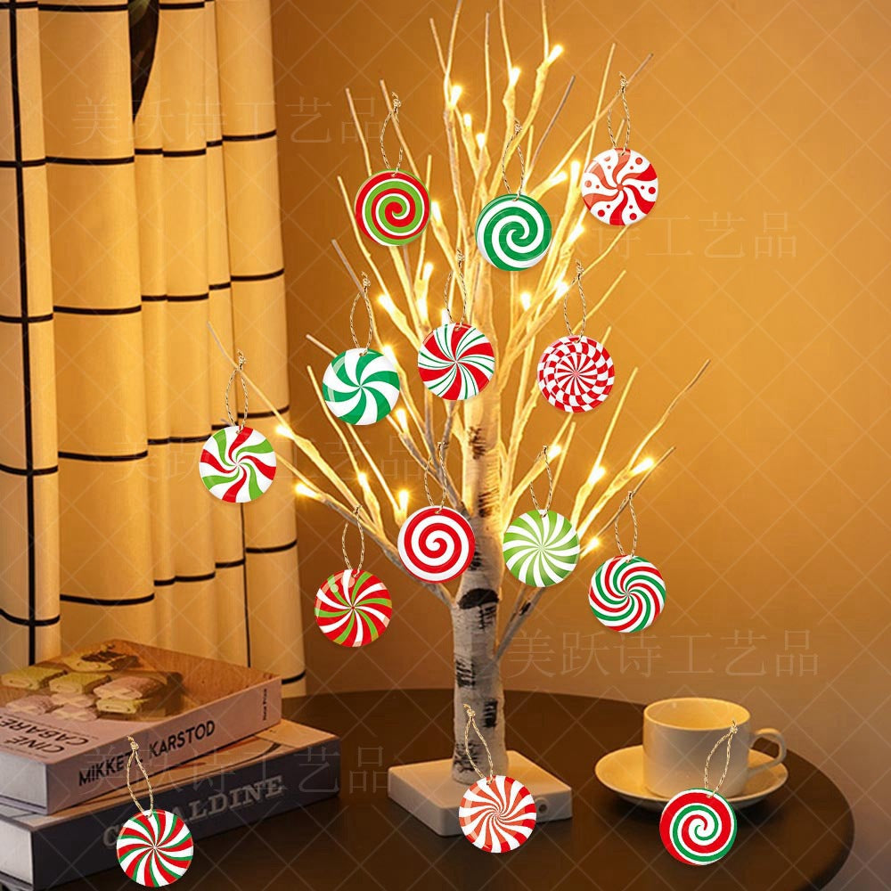 A lit white decorative tree on a table adorned with red and green swirl ornaments, next to stacked books and a white cup, set against a yellow curtain backdrop.