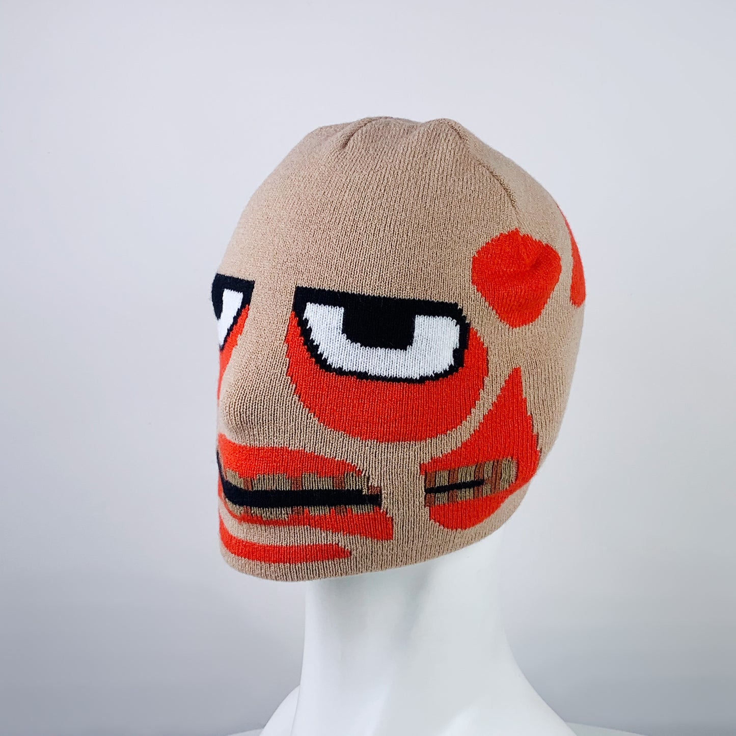 Beige knitted balaclava featuring a stylized face design with bold red and black elements, large cartoonish eyes, and abstract patterns, displayed on a white mannequin head.