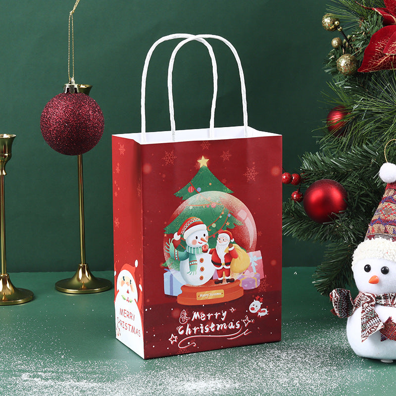 Red Christmas gift bag with snowman and tree design, next to a decorated tree. Includes a sparkling ornament and snowman figure on a green surface lightly dusted with snow.
