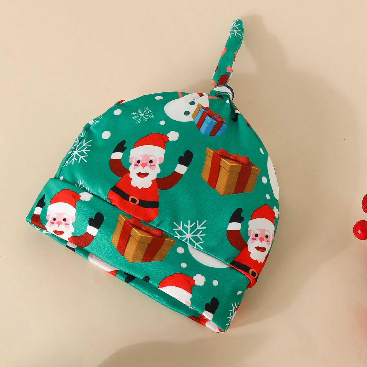 Green baby hat with festive prints of Santa Claus, snowflakes, gifts, and snowmen. The top has a fabric knot. Set against a plain beige background.