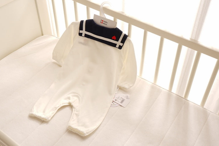 White baby onesie with a navy blue collar and two stripes, adorned with a small red heart, laid on a white mattress, hanging by a coat hanger with tags attached.