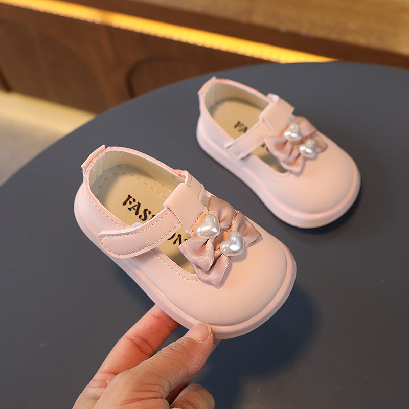 Pink baby shoes with a strap, featuring a decorative bow and heart-shaped pearls on the front. The shoes are placed on a dark surface, with one shoe held up for display.
