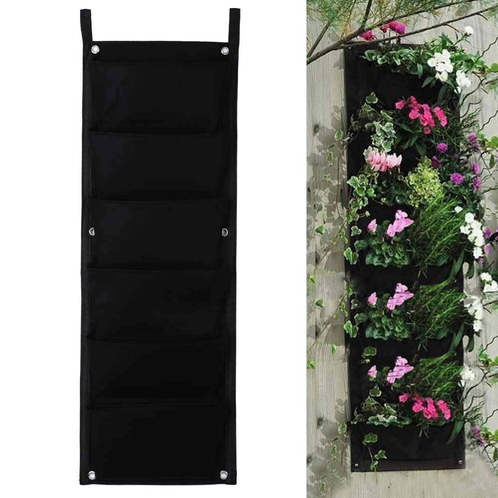 Black vertical gardening pocket with five compartments, shown empty on the left and filled with vibrant pink, white, and purple flowers on the right, mounted on a wooden wall.