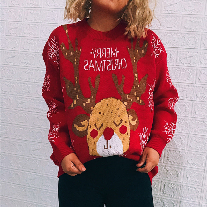 Child in red Christmas sweater featuring a reindeer design and snowflakes, with "Merry Christmas" written. Background is a white brick wall.