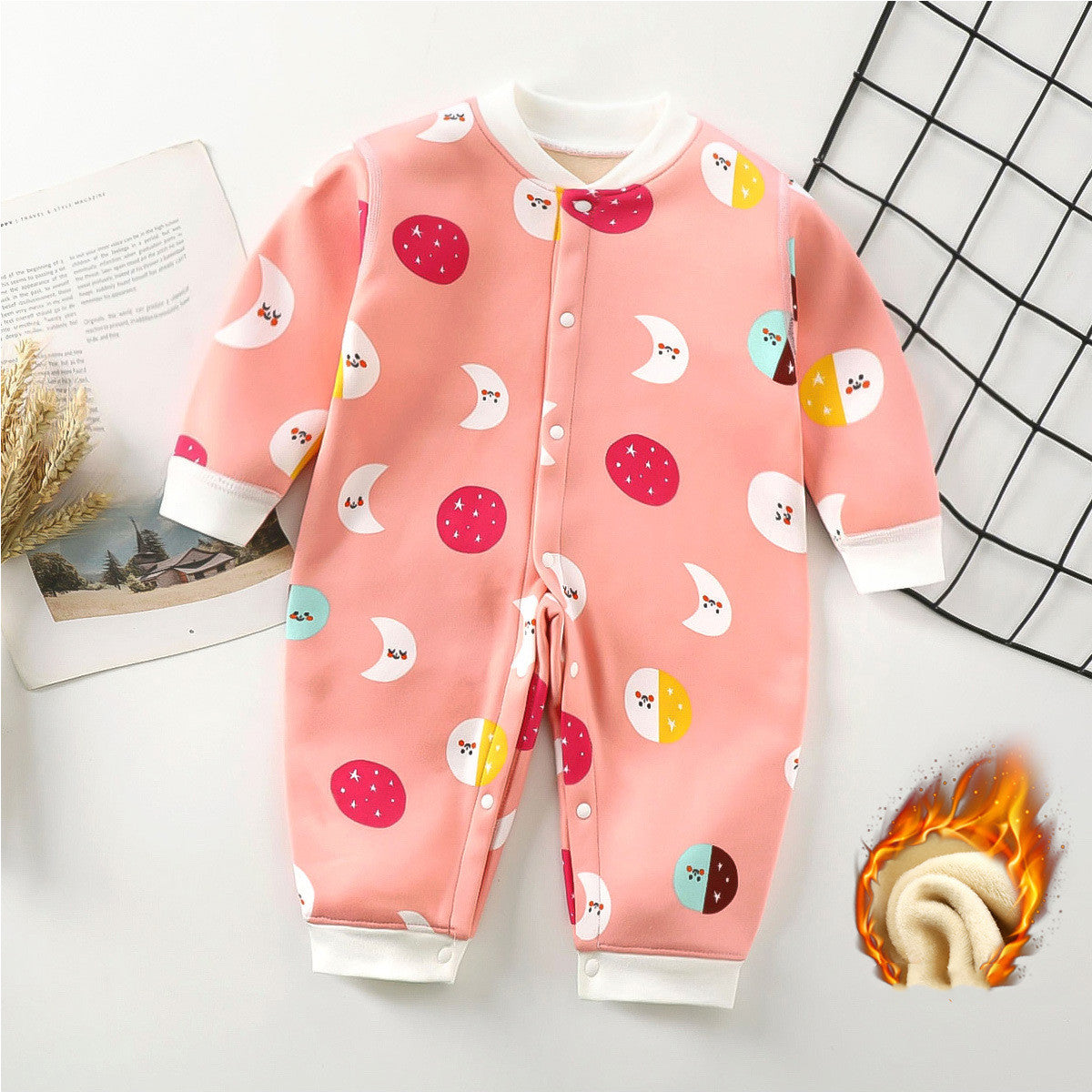 Pink children's jumpsuit with colorful moon and star patterns. Features white cuffs and a button-down front. Beside it, a magazine and decorative wheat on a white table.