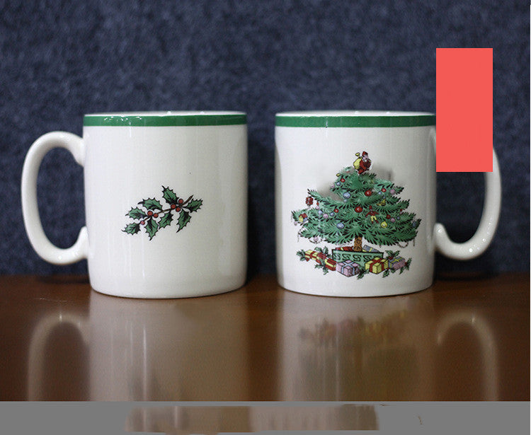 Two white mugs with green rims. The left mug features a holly design, while the right mug showcases a decorated Christmas tree with presents. They are placed on a wooden surface.