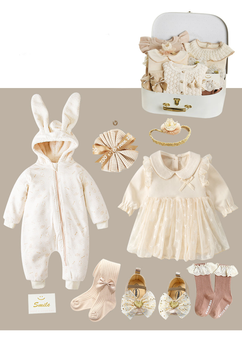 A baby gift set featuring a bunny-ear onesie, cream dress, socks, shoes with bows, and hair accessories, displayed with a suitcase of folded beige baby outfits.