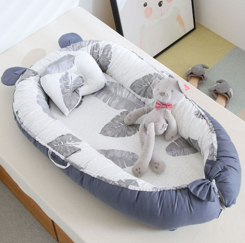 A cozy baby nest with gray and white leaf patterns, featuring a matching pillow and a plush toy cat with a red bow tie. Art and slippers are visible in the background.