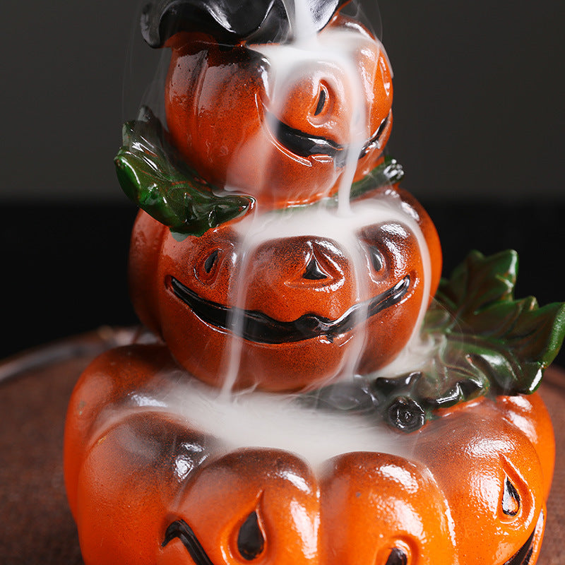 Stacked ceramic pumpkins with jack-o'-lantern faces, spewing white smoke from the top down, creating a whimsical, spooky appearance. Green leaves accent the vibrant orange.