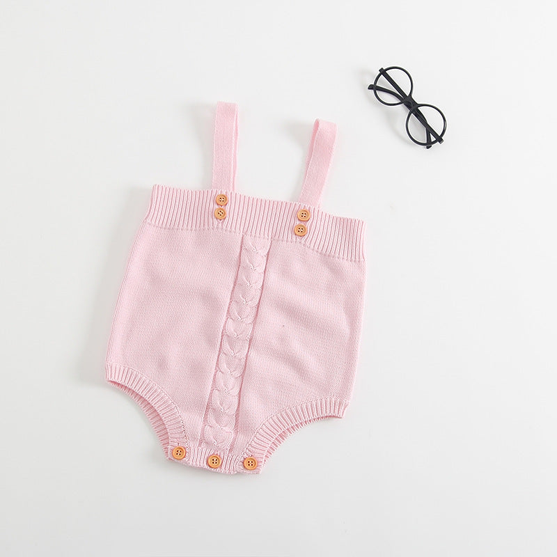 Pink knitted baby romper with wooden buttons and cable pattern detail, laid flat. Features adjustable shoulder straps. Black round glasses are placed nearby on a white background.