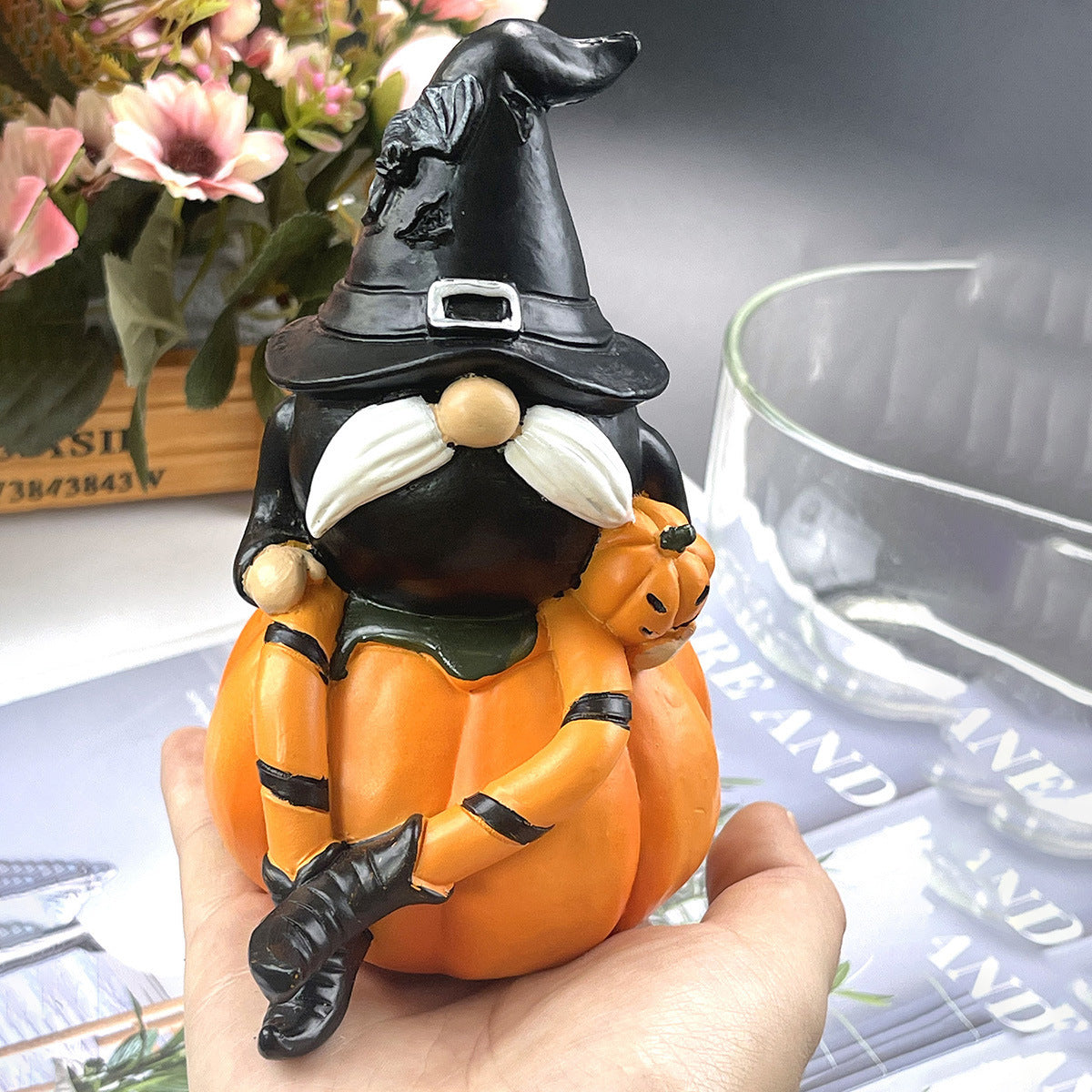 Whimsical gnome figurine in a witch hat, sitting on an orange pumpkin, held in a hand. It features black stripes, white mustache, and a small jack-o'-lantern. Flowers are in the background.