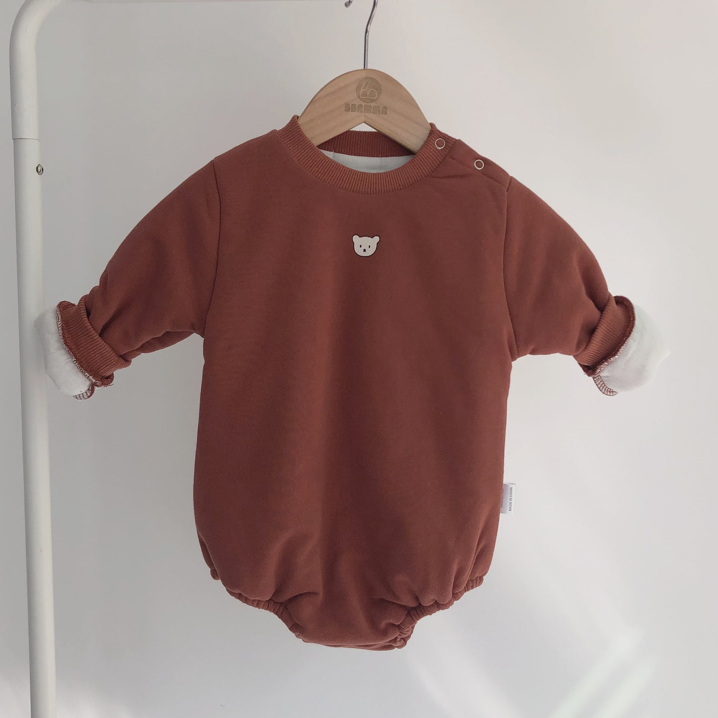 Rust-colored baby romper with long sleeves, cuffed wrists, and a small bear face emblem on the chest. It hangs on a wooden hanger against a light background.