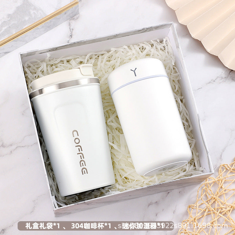 Gift box with a white travel coffee mug and a sleek white cylinder container nestled in shredded paper. The box is set on a marble surface with a beige fan shape nearby.