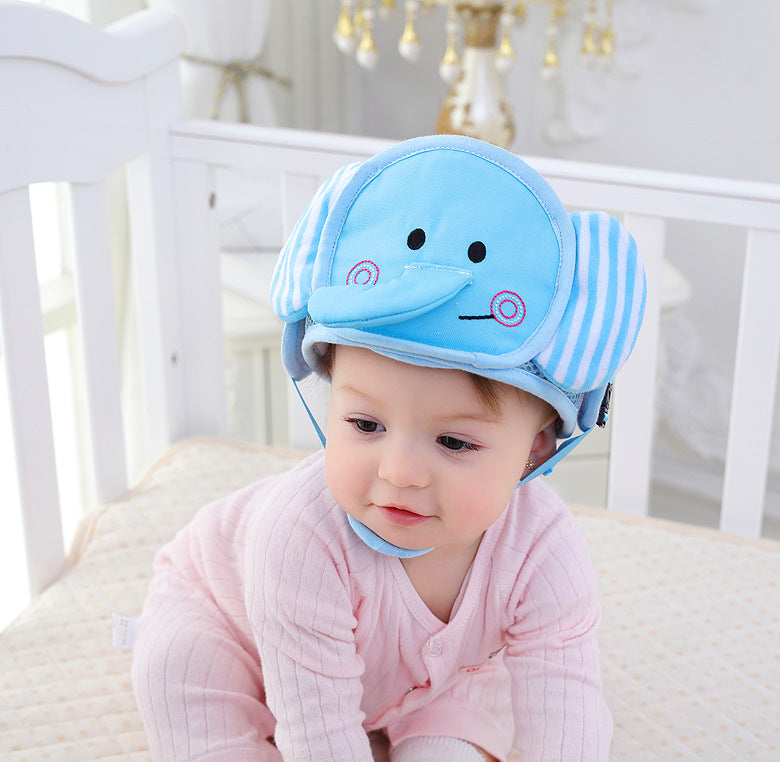 A baby wearing a pink onesie is sitting in a crib, sporting a blue elephant-themed protective helmet. The room has a white and gold color scheme with elegant furnishings.