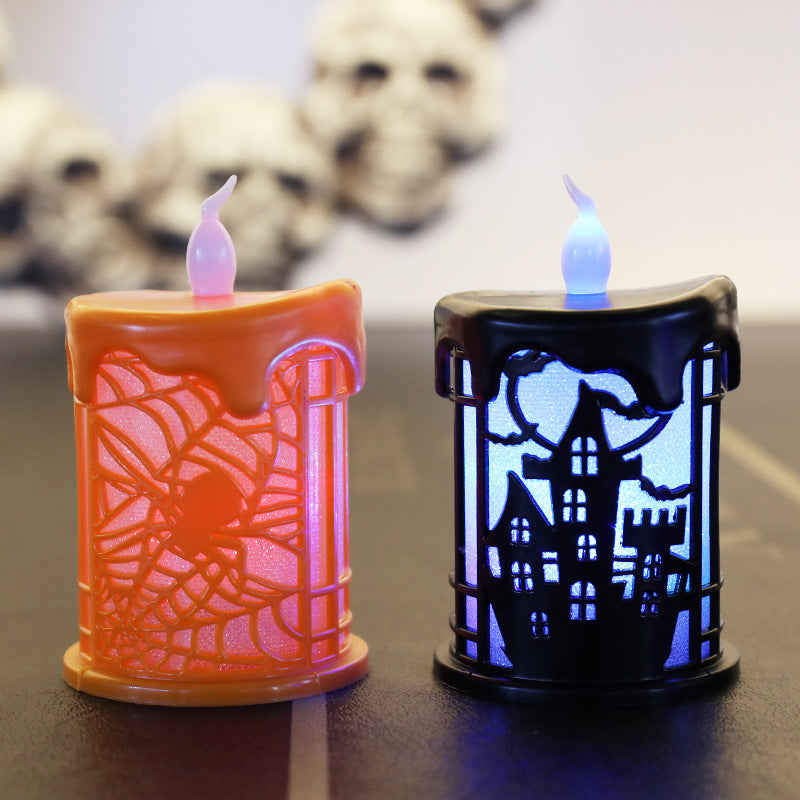 Two decorative LED candles on a table. The orange candle features a spiderweb design with a spider, while the black candle displays a haunted house silhouette against a blue backdrop. Both have melted wax-like tops. Blurred skull decorations are visible in the background.