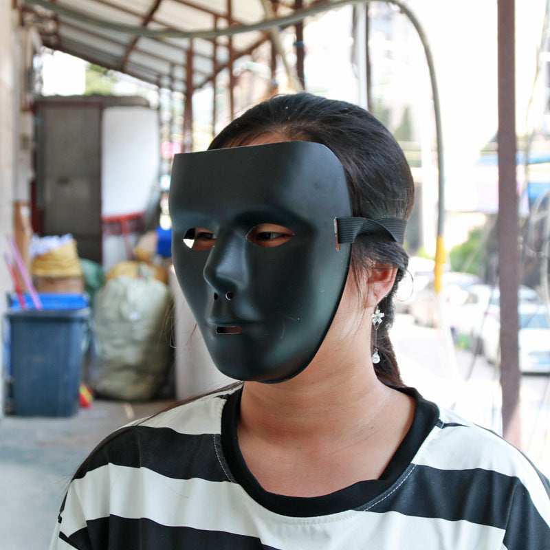A person wearing a black full-face mask stands outdoors. They are dressed in a black and white striped shirt, and their hair is tied back, with silver earrings visible.