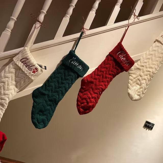 Three knitted Christmas stockings in white, green, and red hang on a staircase railing. Each stocking has a name embroidered on the cuff, with festive holiday decorations.