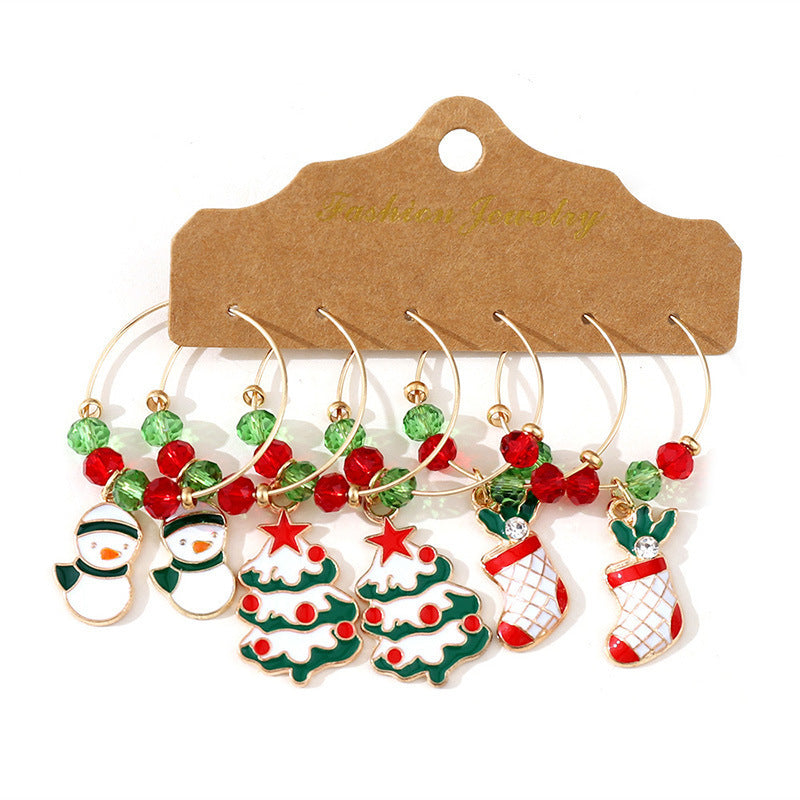 Six gold wine glass charms with festive Christmas designs, including snowmen, trees, and stockings, adorned with red and green beads, on a kraft paper backing.