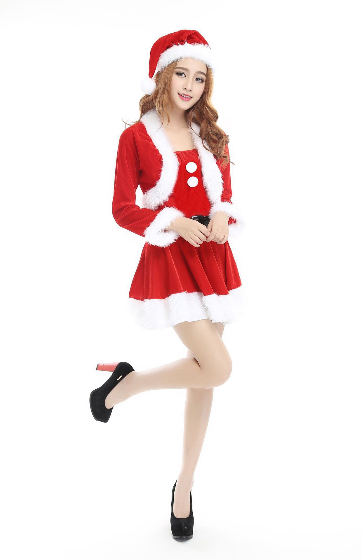 Woman in a festive red and white Santa dress with fur trim and red Santa hat, posing with one leg bent. She wears black high heels and smiles at the camera against a white background.