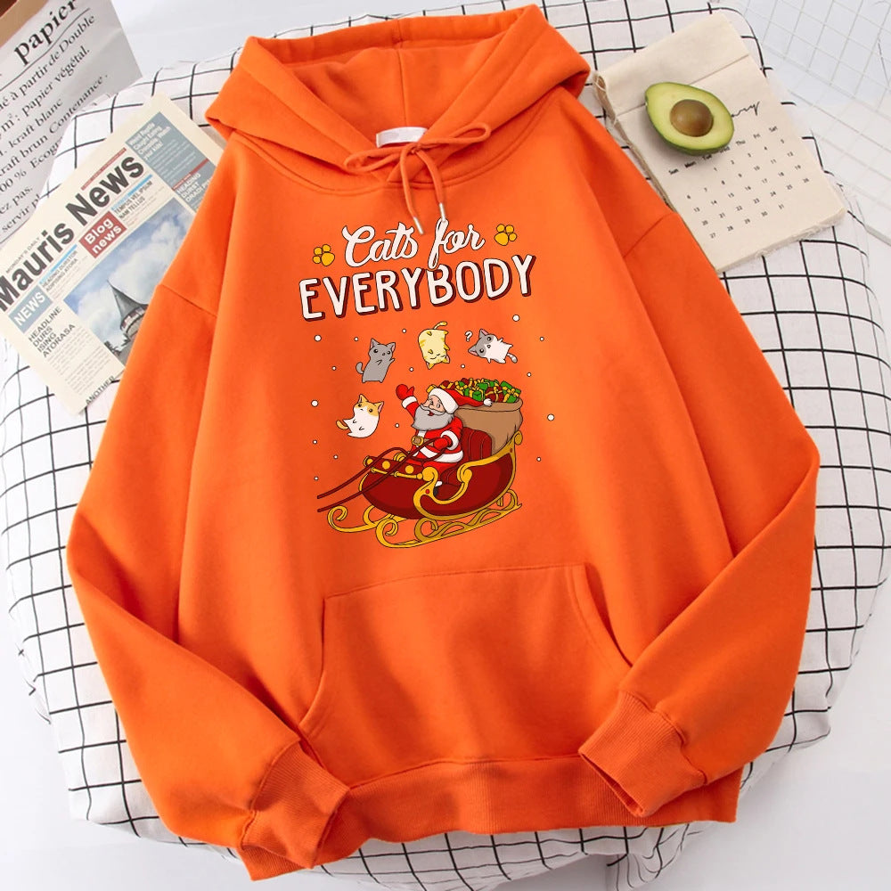 Orange hoodie with "Cats for Everybody" text above a cartoon Santa in a sleigh, surrounded by playful cats. Nearby, there's a newspaper and half an avocado on a tablecloth.