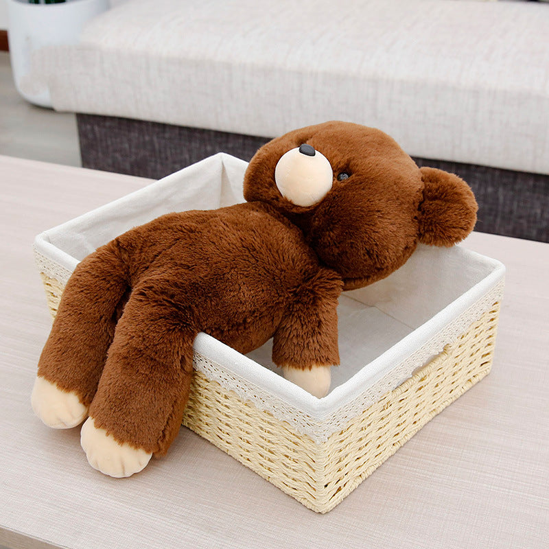 A plush brown teddy bear is lying on its back, partially inside a woven basket with white lining, placed on a light wood table in a cozy room with a couch in the background.