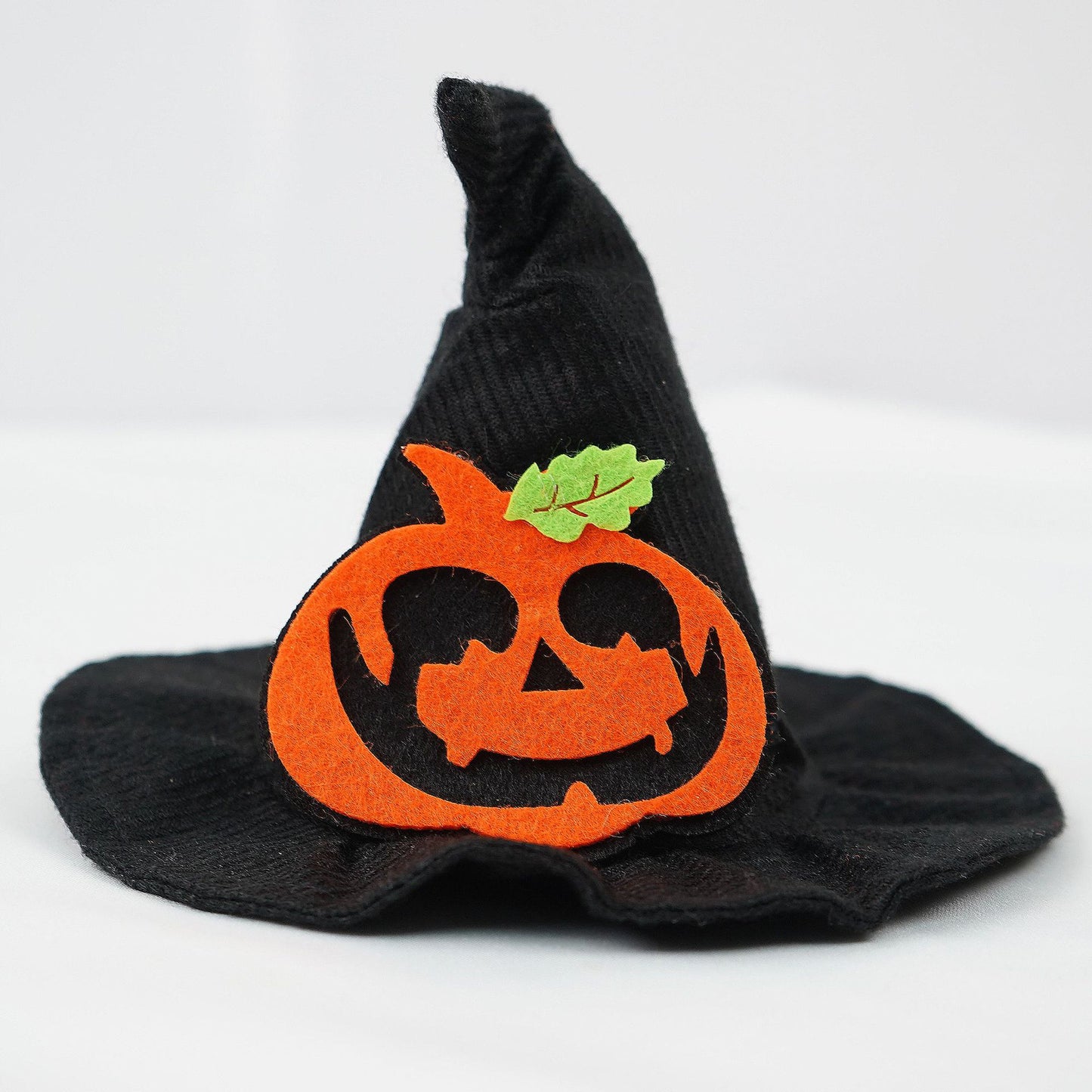 A small black witch hat adorned with a smiling orange felt jack-o'-lantern and a green leaf. Perfect for Halloween decorations or festive attire.