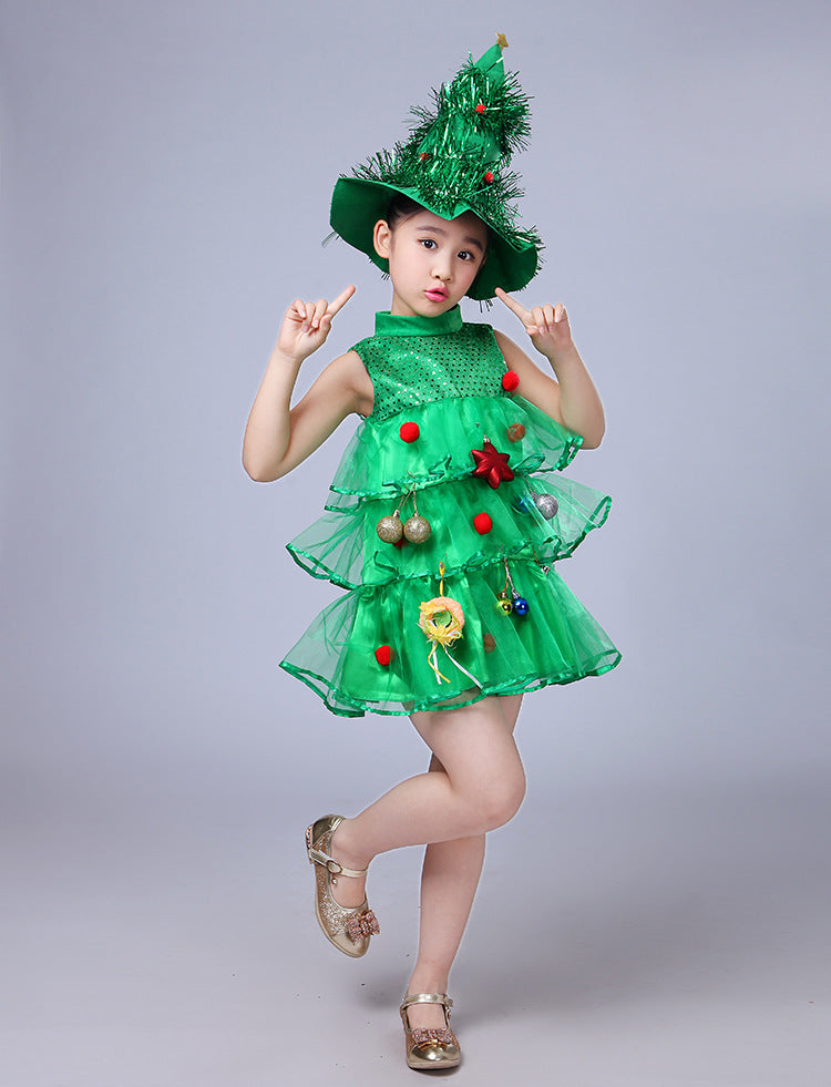 Child dressed as a Christmas tree, wearing a green tulle dress with ornaments and a matching hat adorned with tinsel, striking a playful pose against a gray background.