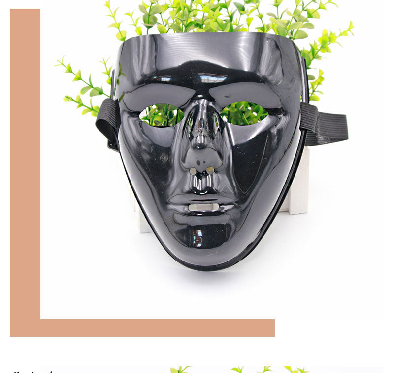 A shiny black face mask with a neutral expression, positioned in front of a potted plant, with green leaves visible around the mask, against a simple white background.