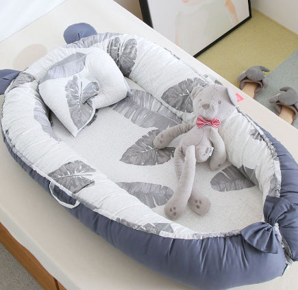 A soft, gray and white leaf-patterned baby nest with a matching pillow and gray plush cat toy with a red bow tie resting inside.