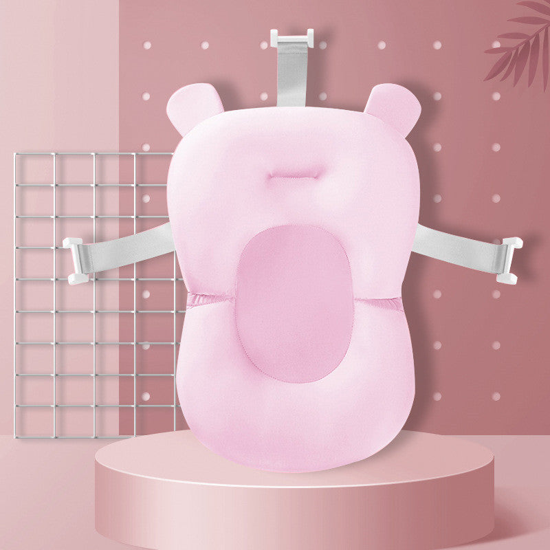 Pink baby bath cushion with headrest and securing straps, featuring bear-like ears. Displayed on a round platform against a pink polka-dot and grid-patterned background.
