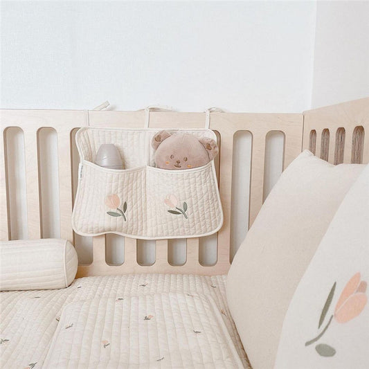 Crib with quilted bedding and a soft toy bear. A quilted organizer with tulip embroidery hangs on the rail, holding a bottle and the toy. Neutral tones create a calming vibe.