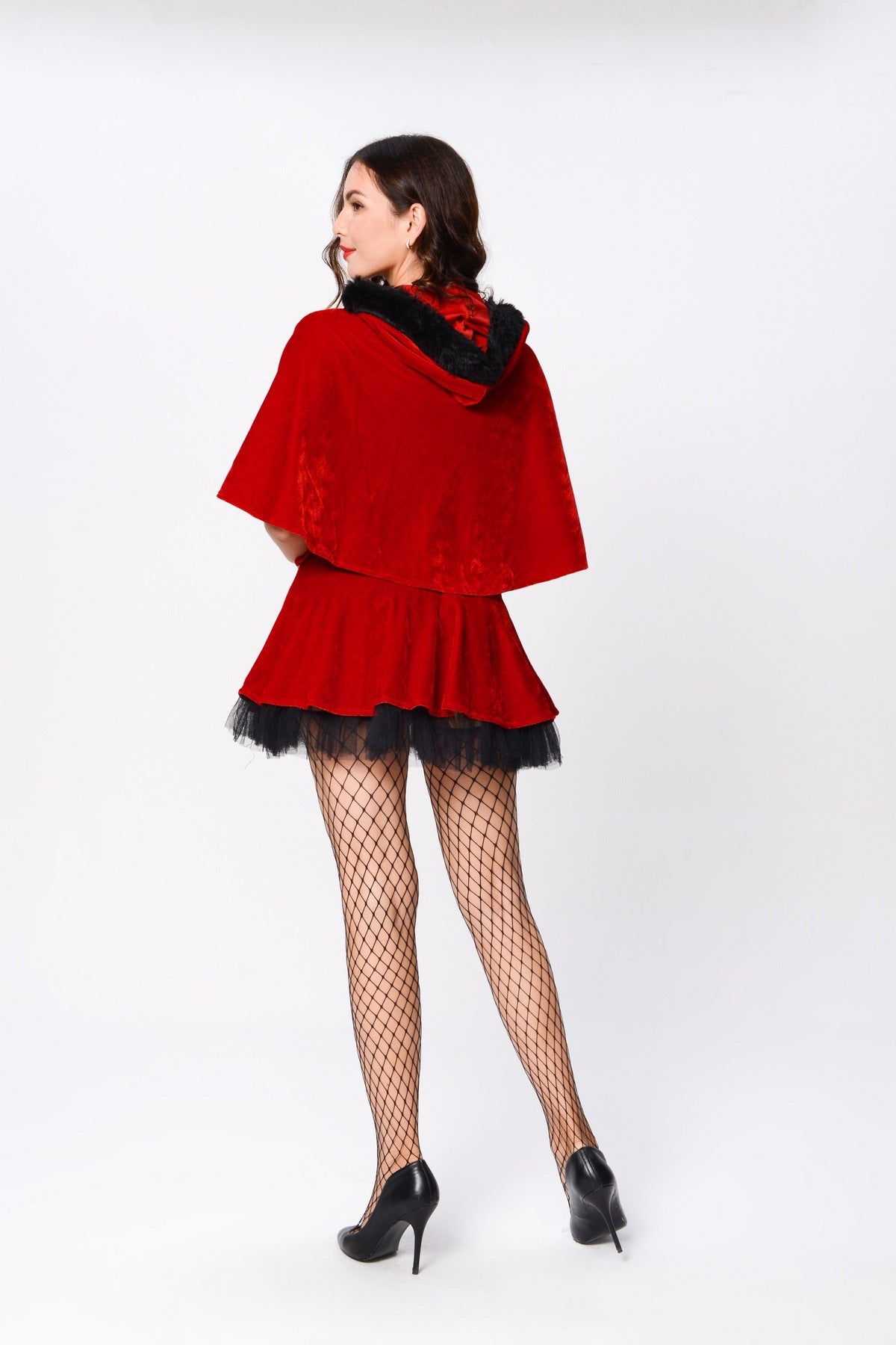 Woman in a red hooded cape and skirt, with black detailing, fishnet stockings, and black heels, standing against a white background, facing slightly away from the camera.