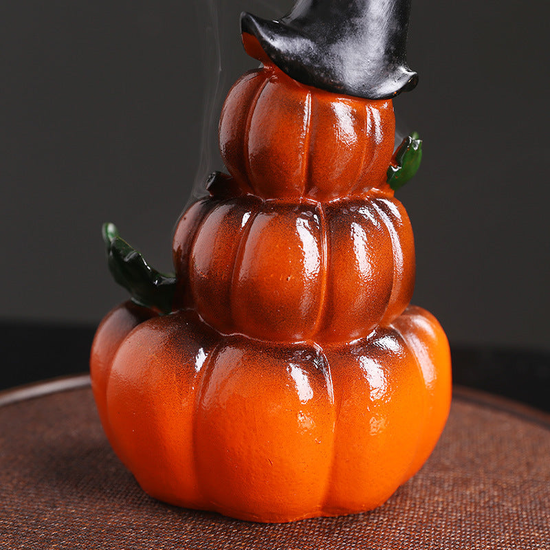 A glossy, stacked ceramic pumpkin ornament with three tiers, topped with a black witch hat. The piece includes small green leaves, set against a dark background.