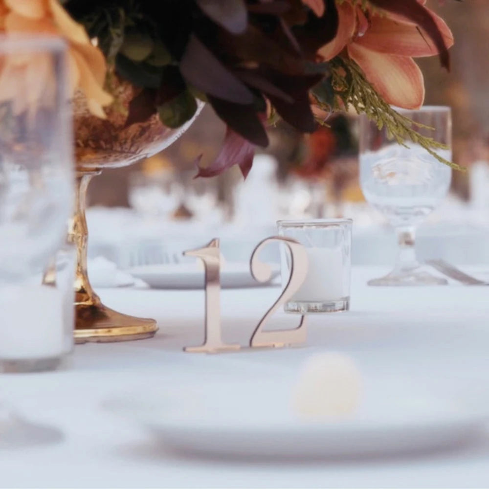 Elegant table setting with a metallic number 12 centerpiece, surrounded by white candles and floral arrangements, on a white tablecloth with glassware in the background.