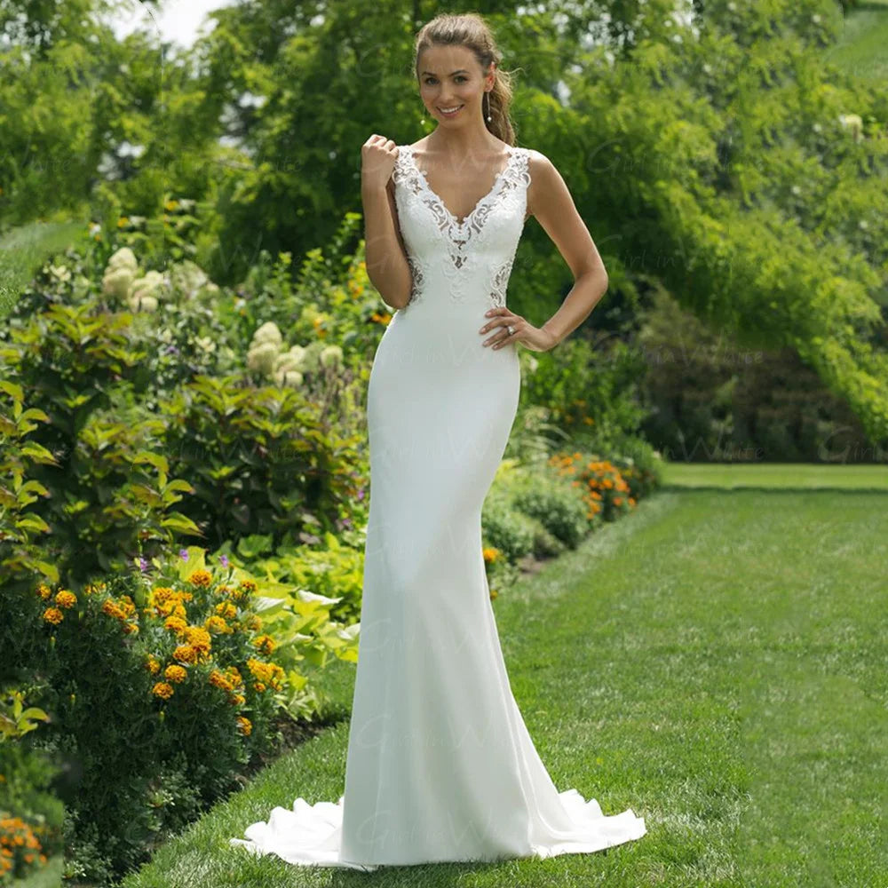 Bride in a sleeveless, lace-detail wedding dress stands on a lush garden path, surrounded by green foliage and colorful flowers, smiling and holding her dress.