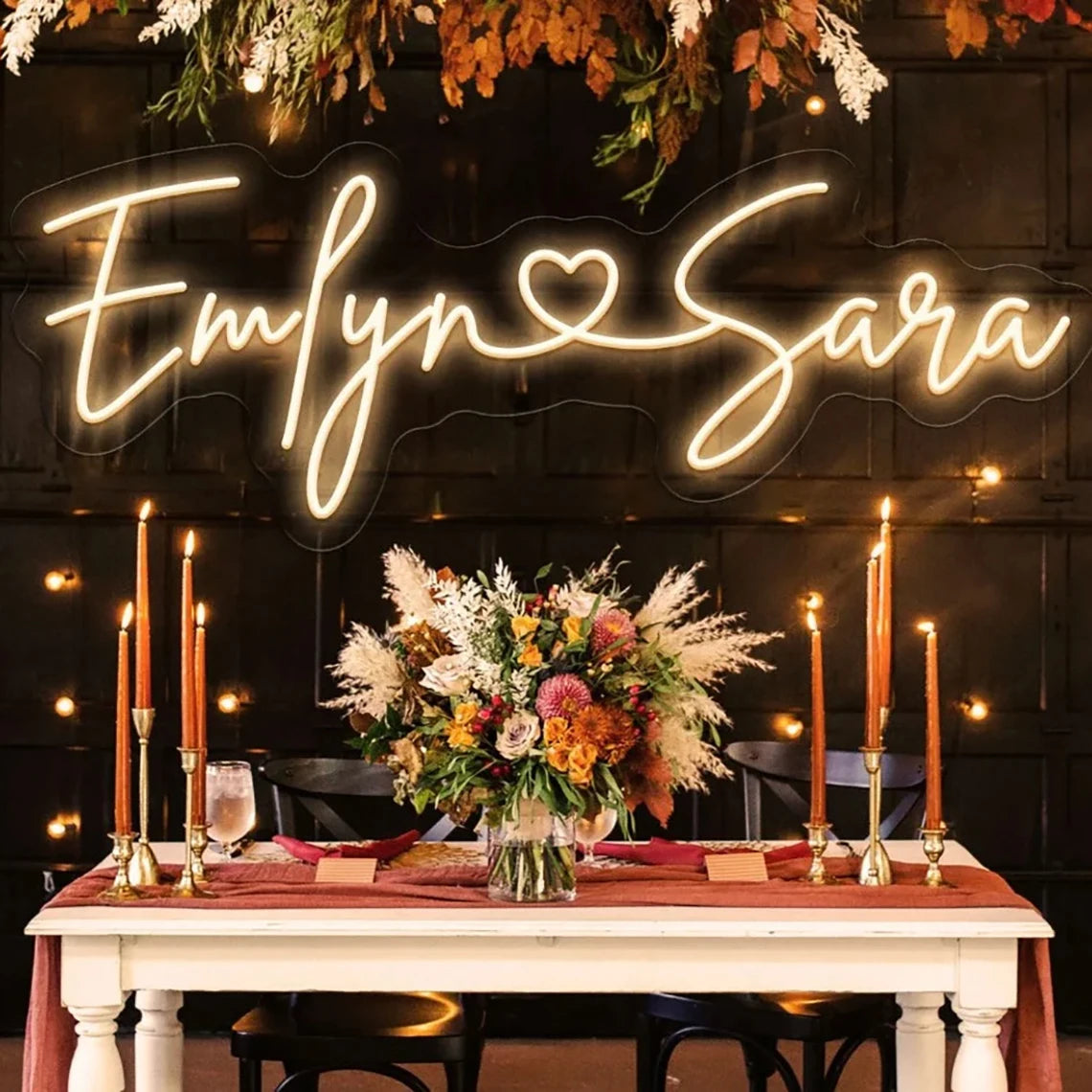 A chic table setup with a vibrant floral centerpiece and tall candles, beneath a warm neon sign displaying intertwined names. The background has foliage and subtle string lights.