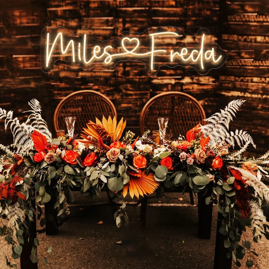 A rustic table decorated with vibrant orange and pink flowers, green foliage, and pampas grass. Behind, "Miles ♥ Freda" glows in neon against a wooden wall.