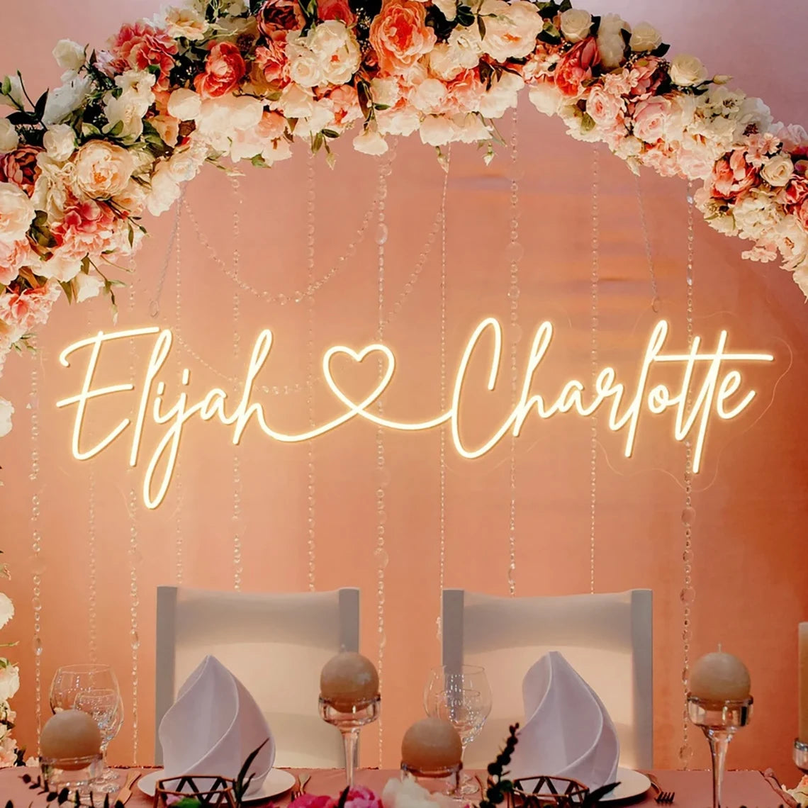 Neon sign reads "Elijah ♥ Charlotte" against a pink backdrop adorned with a floral arch of peach and white roses. Elegant table setting with folded napkins and glassware below.