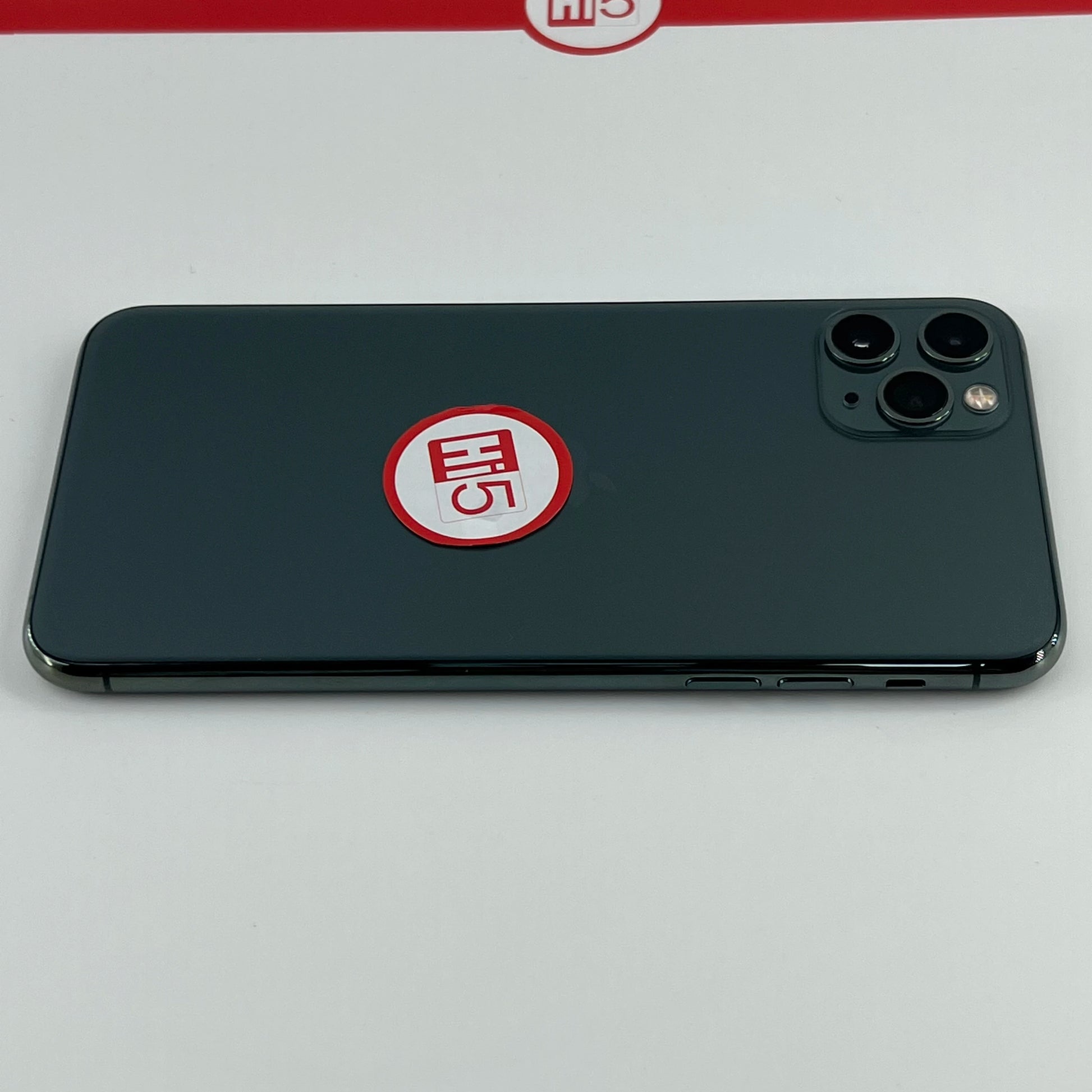 A smartphone with a dark, sleek back lies on a surface. A red and white sticker with "Hi5" is placed near the triple camera lens. A similar logo appears on the backdrop.