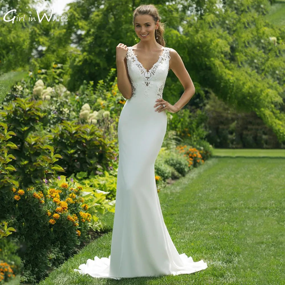Bride in an elegant, form-fitting wedding dress with lace details, standing on a garden path. Lush greenery and colorful flowers surround her under a bright, clear sky.