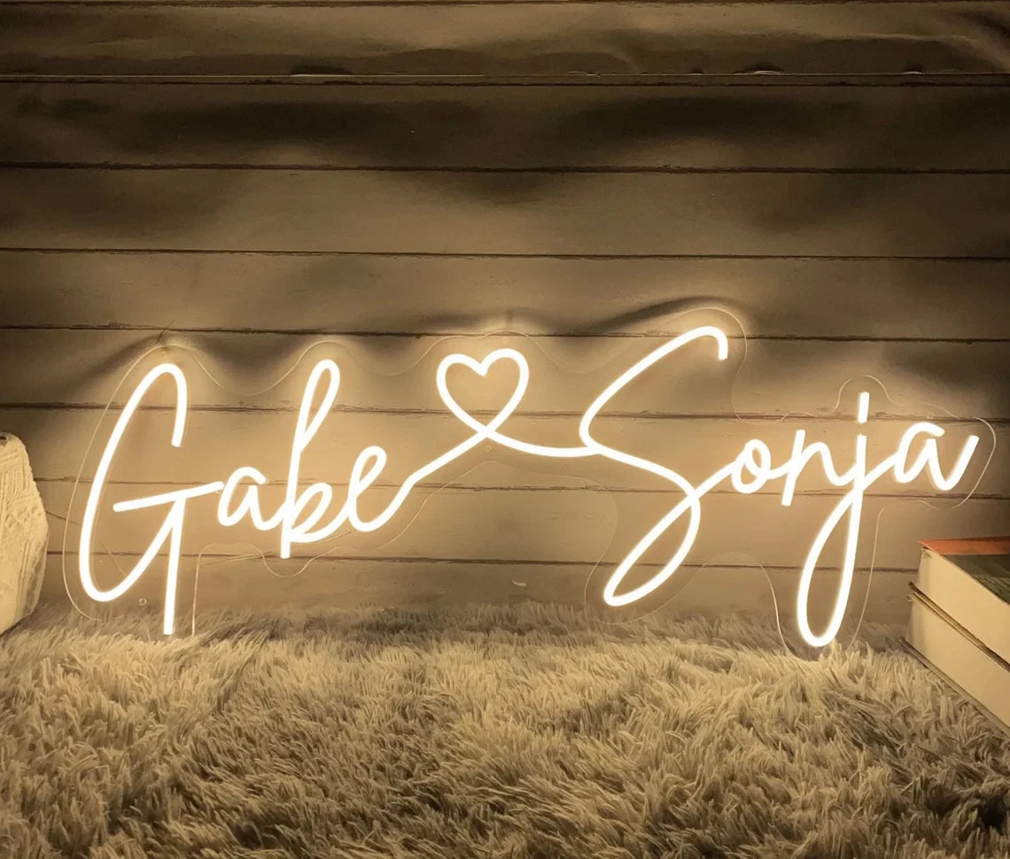 Illuminated cursive neon sign reading "Gabe ♥ Sonja," set against a wooden backdrop. The sign emits a warm glow on the surrounding gray, shaggy rug.